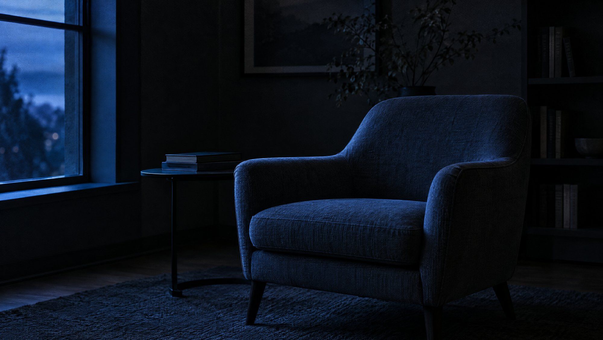 An empty upholstered armchair beside a window in a dark reading room, in soft blue evening light.