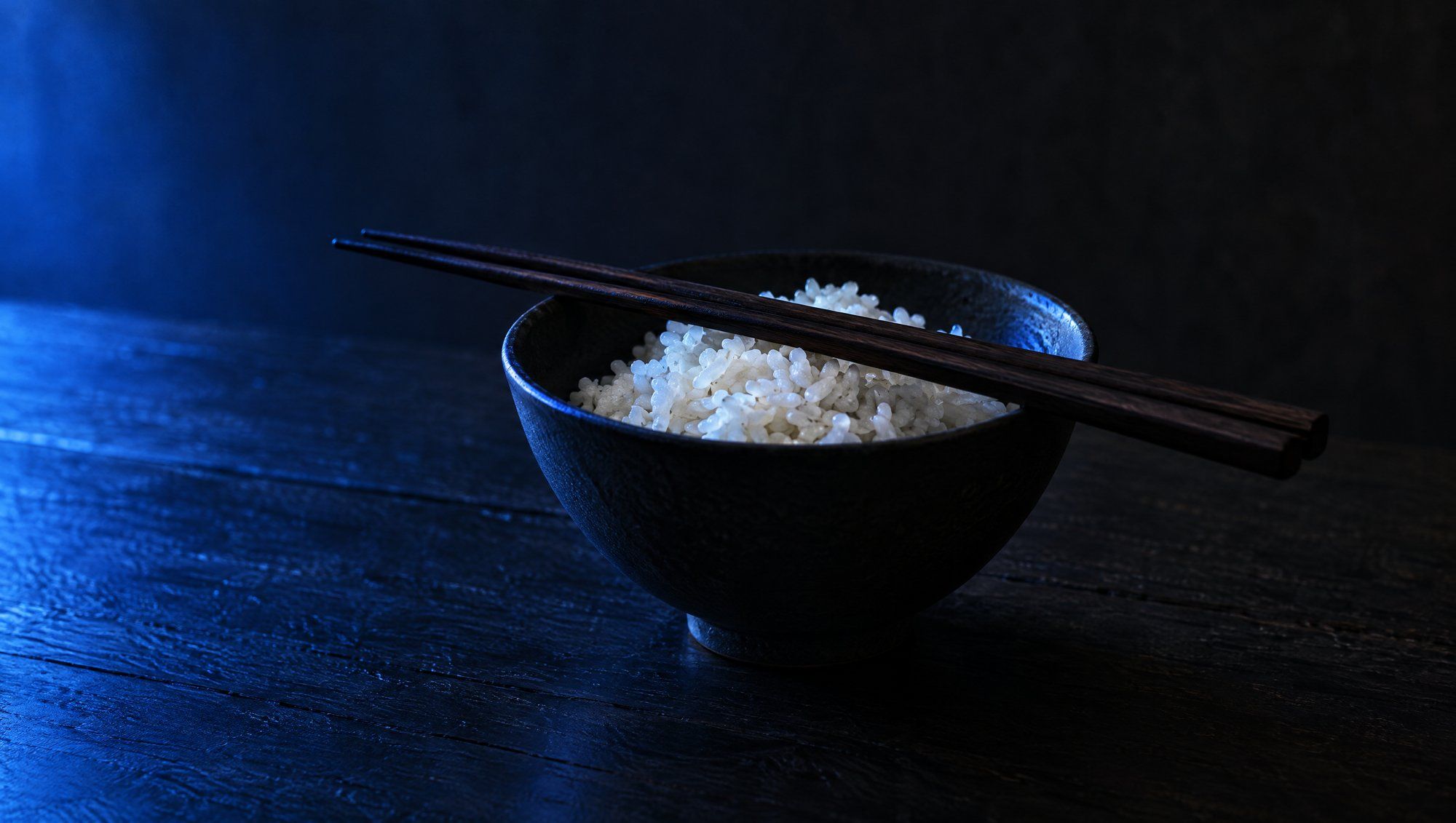 A dark ceramic bowl of white rice with wooden chopsticks laid across it on a dark wooden table, lit from the side in blue.