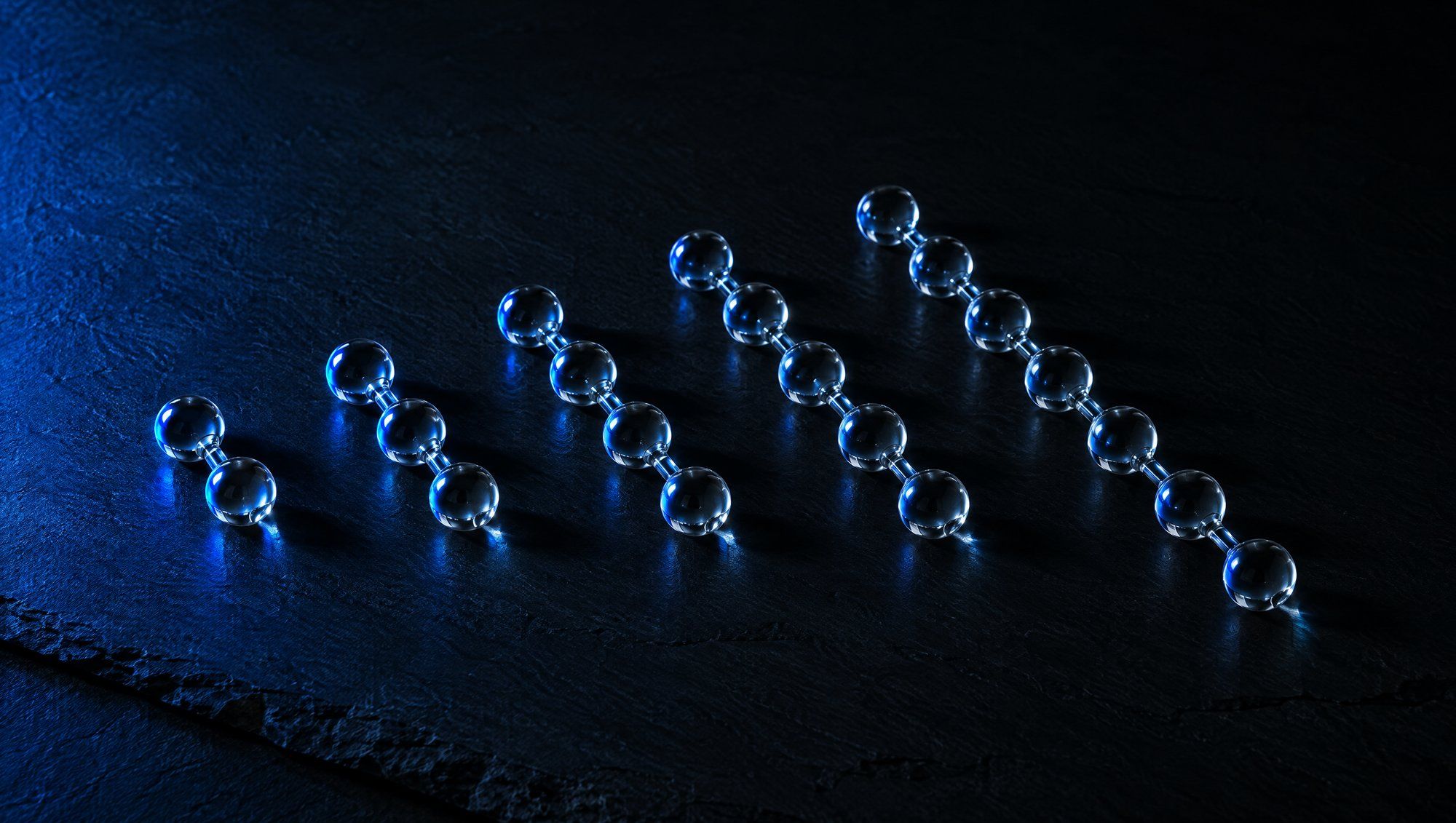 Five chains of clear glass beads of increasing length lying in a diagonal row on dark slate, lit from the left by a navy-blue light.