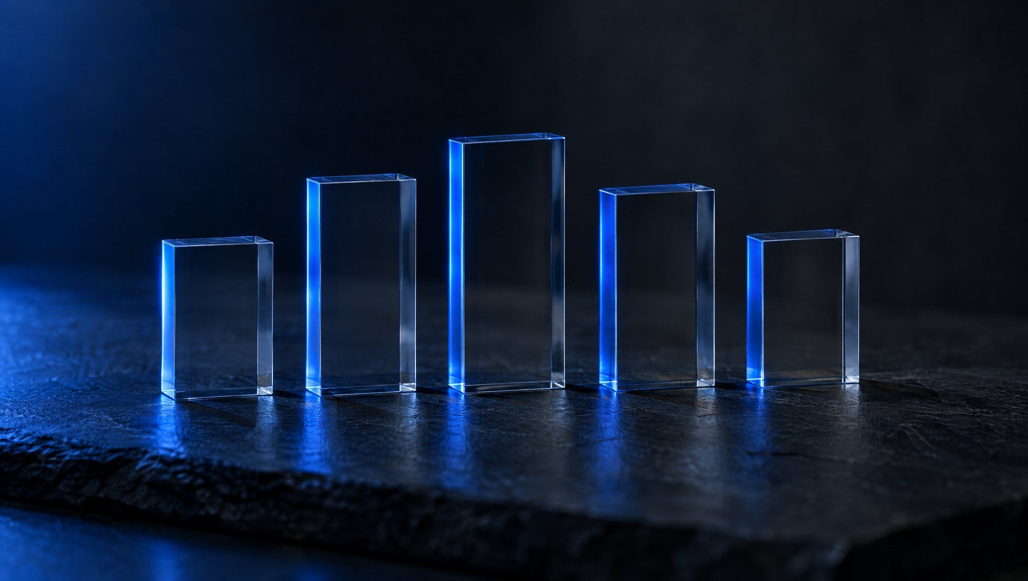Five clear glass blocks of different heights standing in a row on dark slate, each edge lit by a thin line of navy-blue light.
