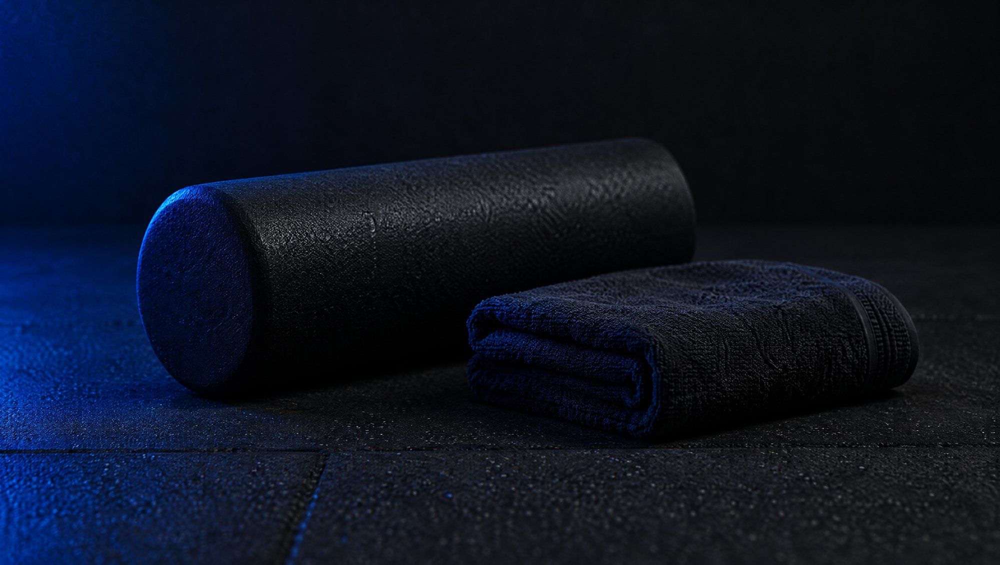 A black foam roller and a folded dark towel on a dark rubber gym floor, lit from one side in blue.