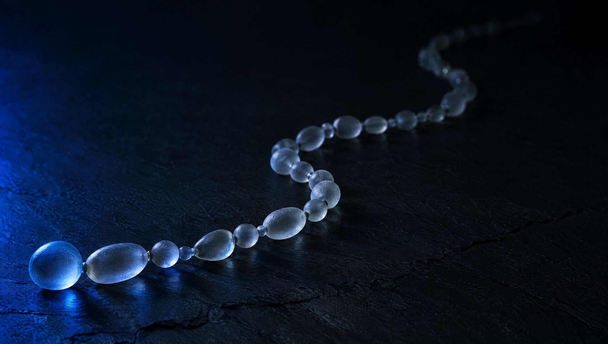 A long chain of frosted glass beads of varied shapes curving across dark slate, the near end lit in navy-blue light and the far end fading into shadow.
