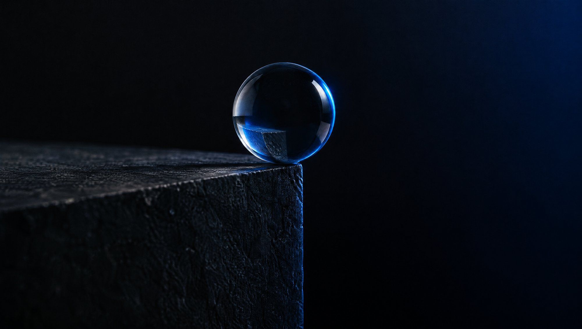 A clear glass sphere resting at the very edge of a dark stone block, in navy-blue rim light with deep shadow beyond.