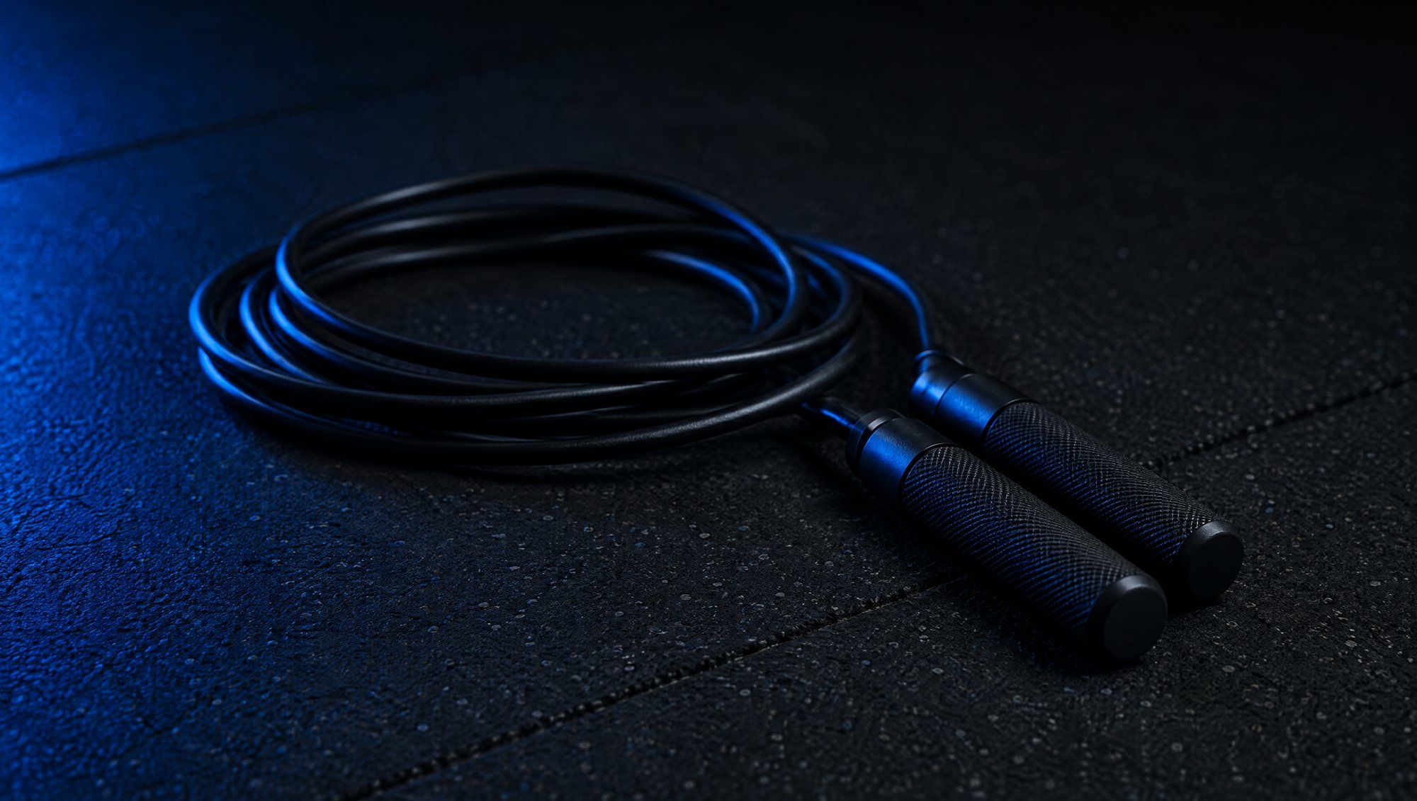 A black jump rope coiled on a dark rubber gym floor, its handles resting together, lit from the side in blue.