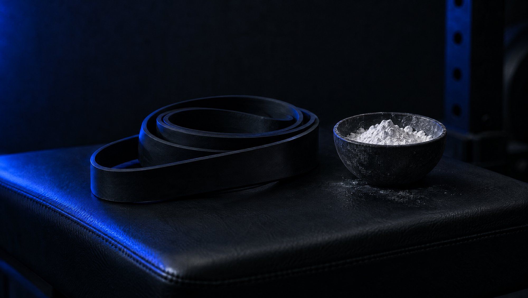 A coiled black resistance band beside a small bowl of white lifting chalk on a dark gym bench, in navy-blue rim light.