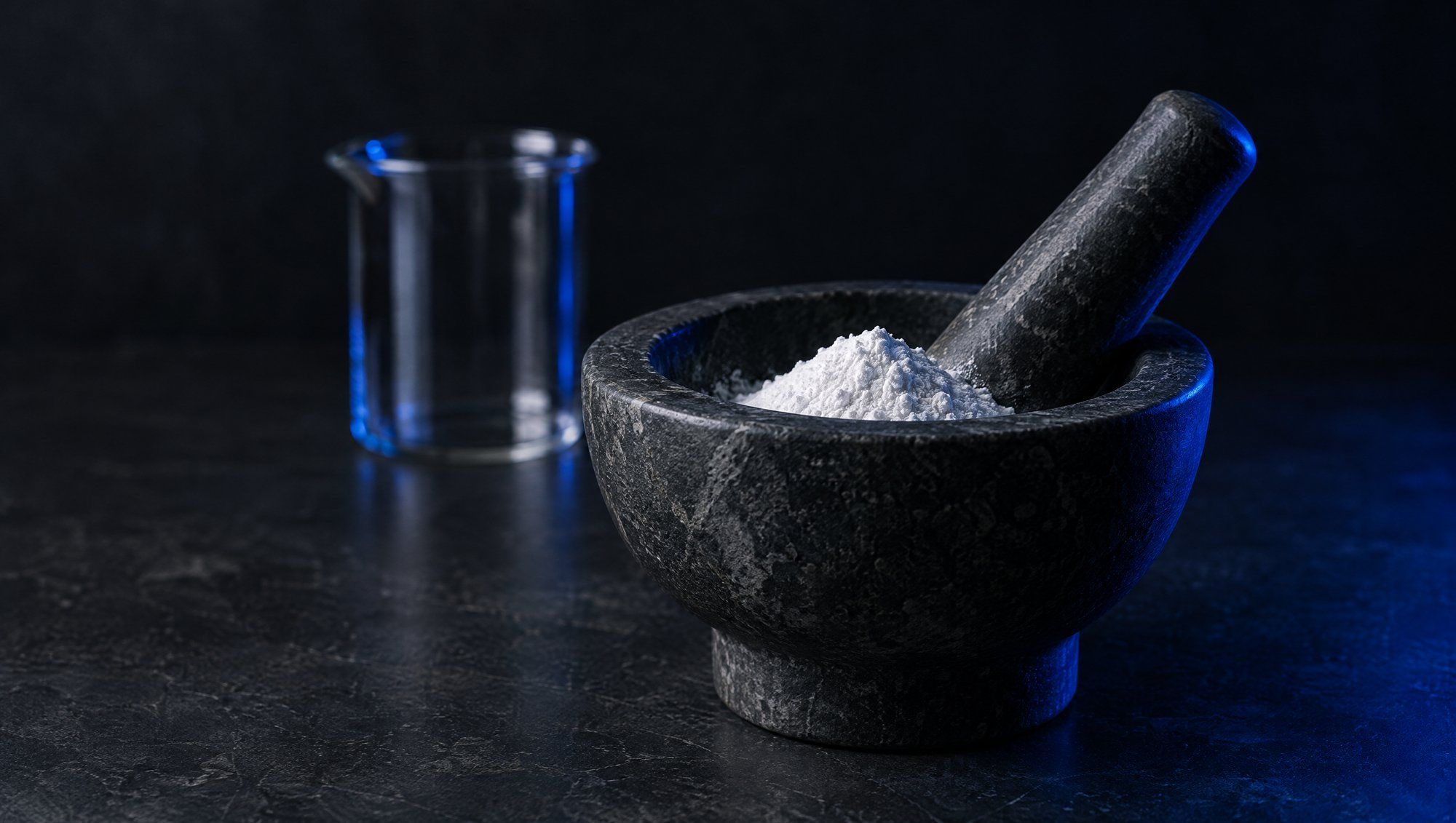 A dark stone mortar and pestle holding a small mound of white powder, an empty glass beaker behind it, on a dark workbench.