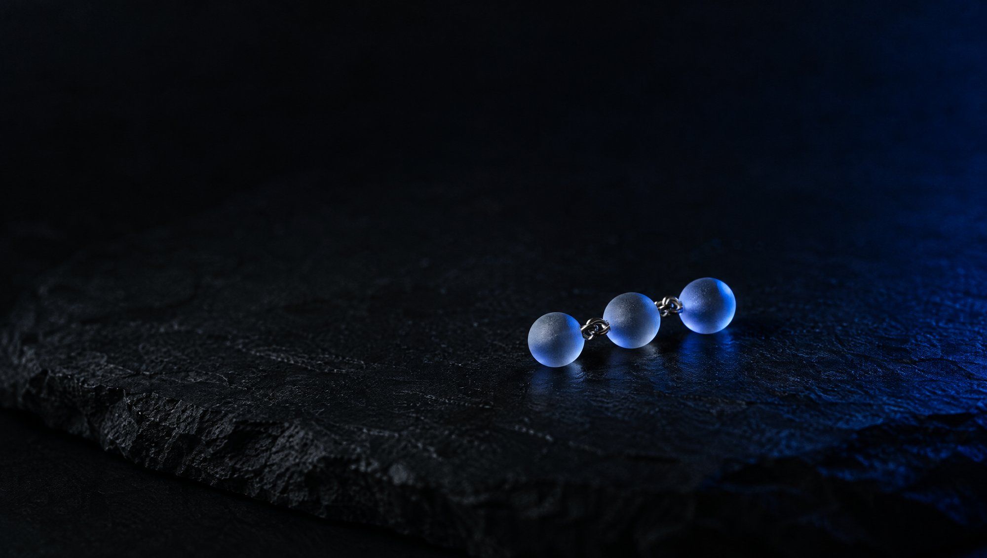 A tiny chain of three frosted glass beads on the edge of a dark slate slab, in navy-blue rim light.