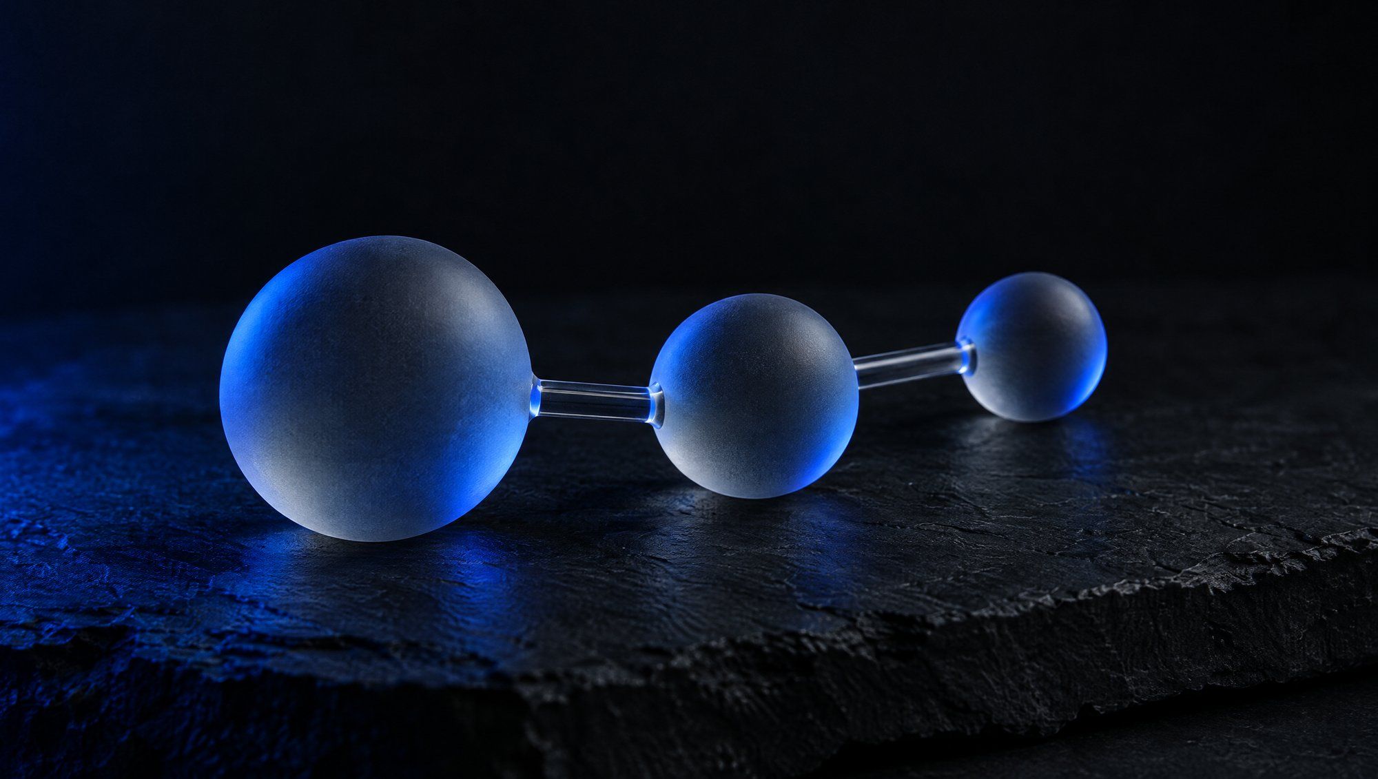Three frosted glass spheres of decreasing size joined by short glass rods, resting on dark slate in navy-blue light.
